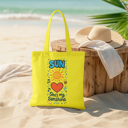  Yellow tote bag with a sun, heart, and "She's my Sunshine" text on a sandy beach with a straw hat and white towel on a wooden stool in the background.