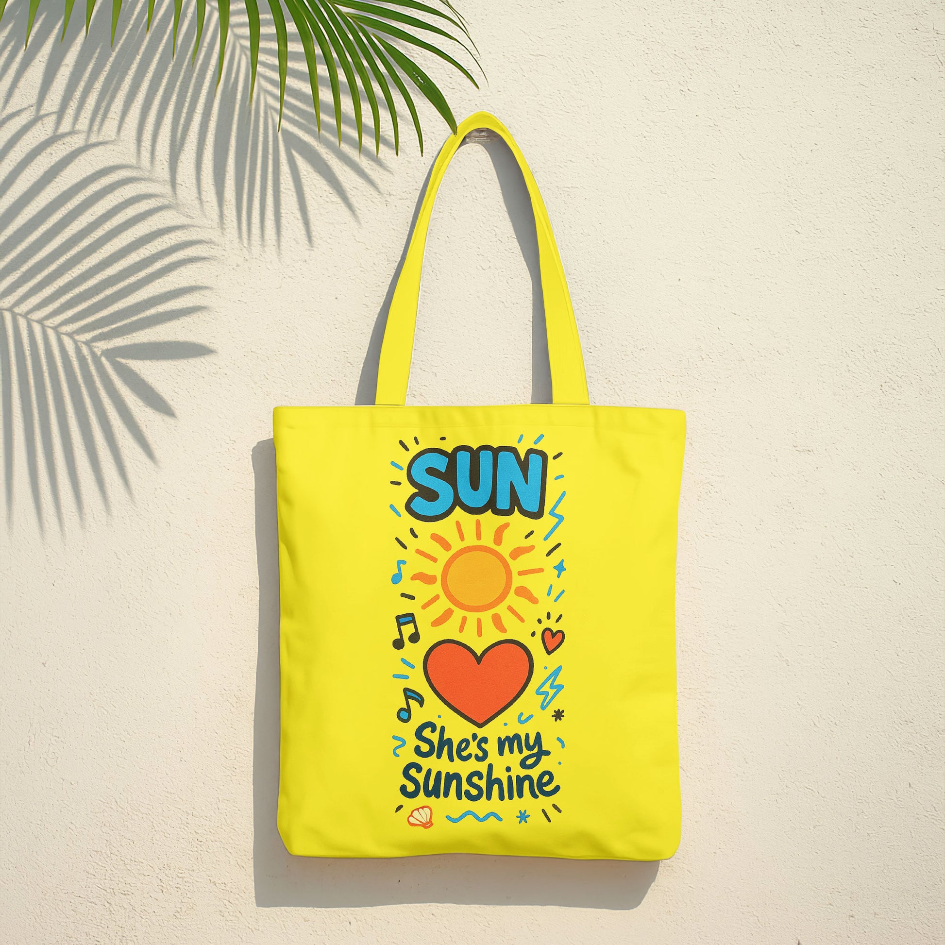  Yellow tote bag with a sun, heart, and "She's my Sunshine" text on a sandy beach with a straw hat and white towel on a wooden stool in the background.