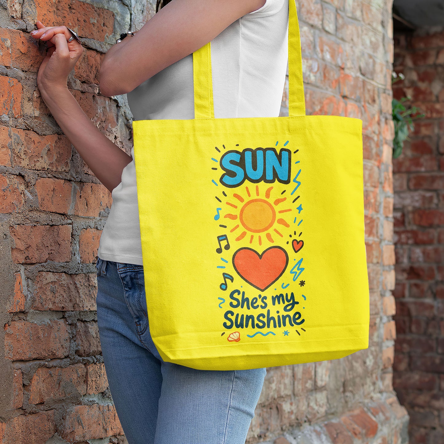  Yellow tote bag with a sun, heart, and "She's my Sunshine" text on a sandy beach with a straw hat and white towel on a wooden stool in the background.