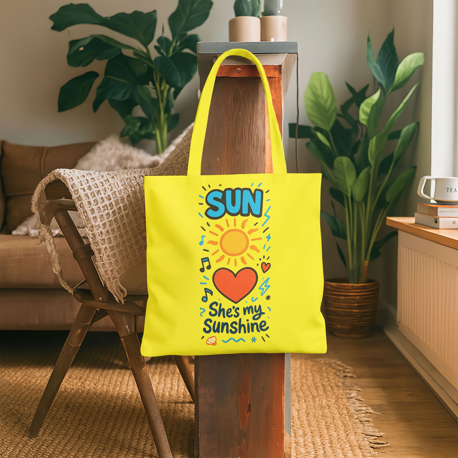  Yellow tote bag with a sun, heart, and "She's my Sunshine" text on a sandy beach with a straw hat and white towel on a wooden stool in the background.