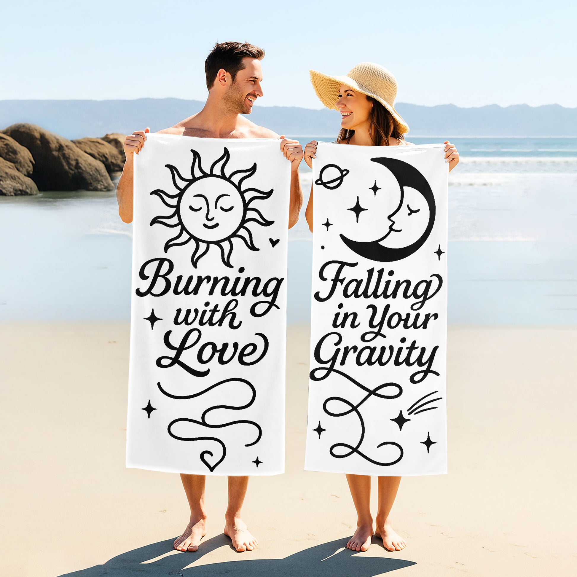 Two people holding up custom white beach towels with black sun and moon designs and romantic phrases, "Burning with Love" and "Falling in Your Gravity," on a sunny beach.