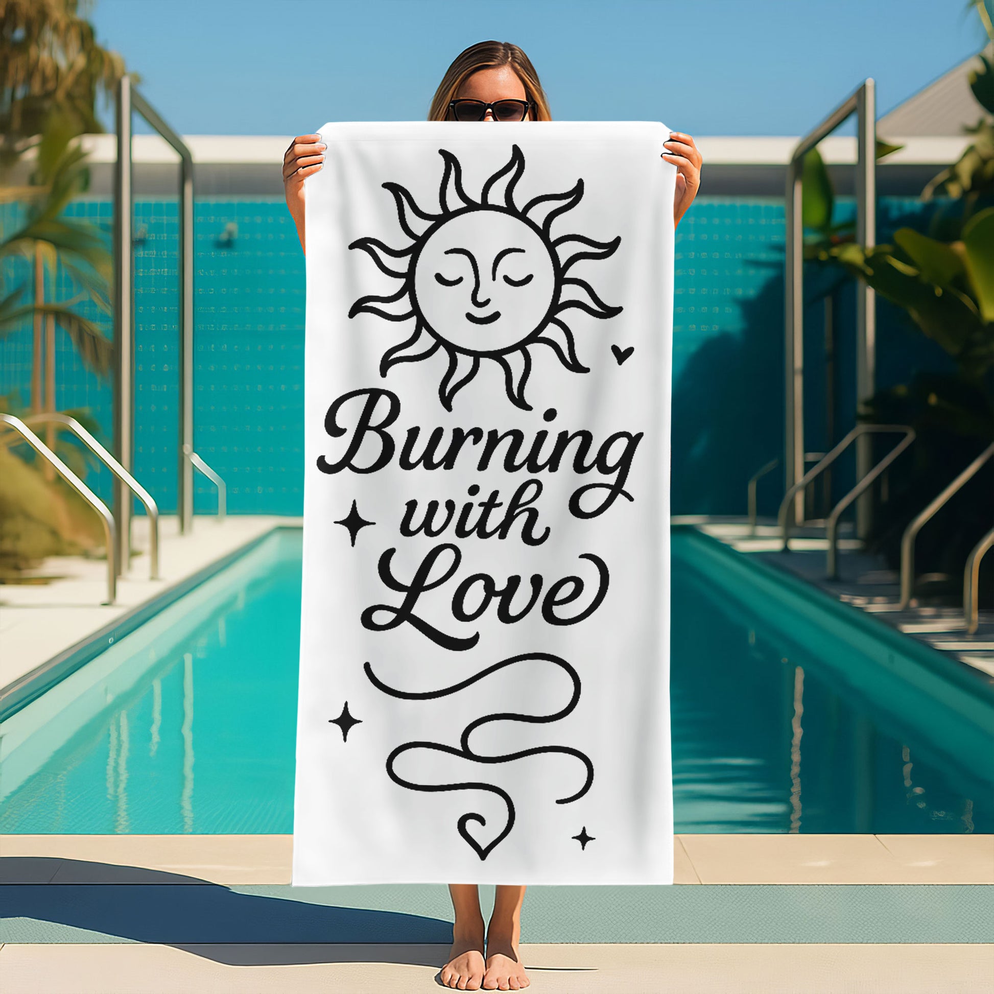 Two people holding up custom white beach towels with black sun and moon designs and romantic phrases, "Burning with Love" and "Falling in Your Gravity," on a sunny beach.