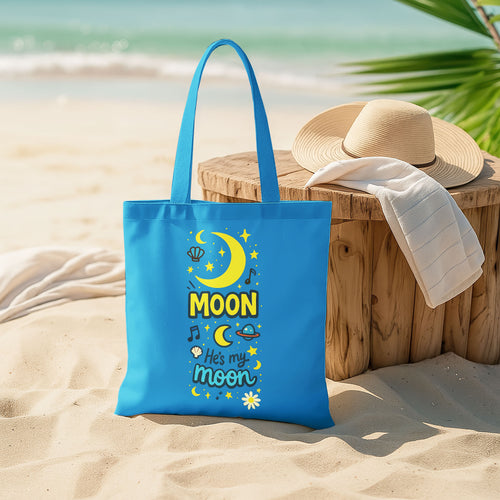 Blue tote bag with yellow moon, stars, and "He's my moon" text on a sandy beach with a straw hat and towel in the background.