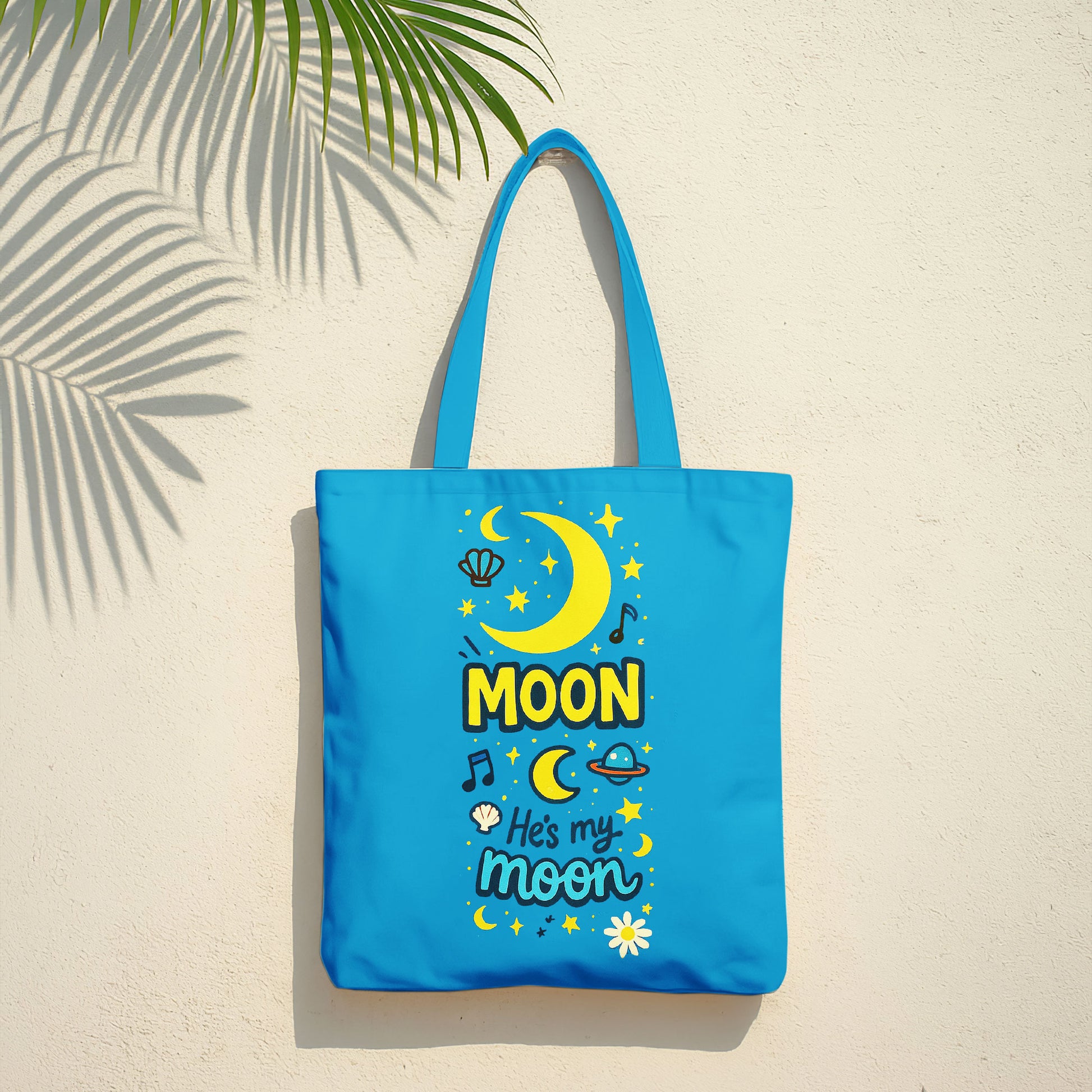Blue tote bag with yellow moon, stars, and "He's my moon" text on a sandy beach with a straw hat and towel in the background.