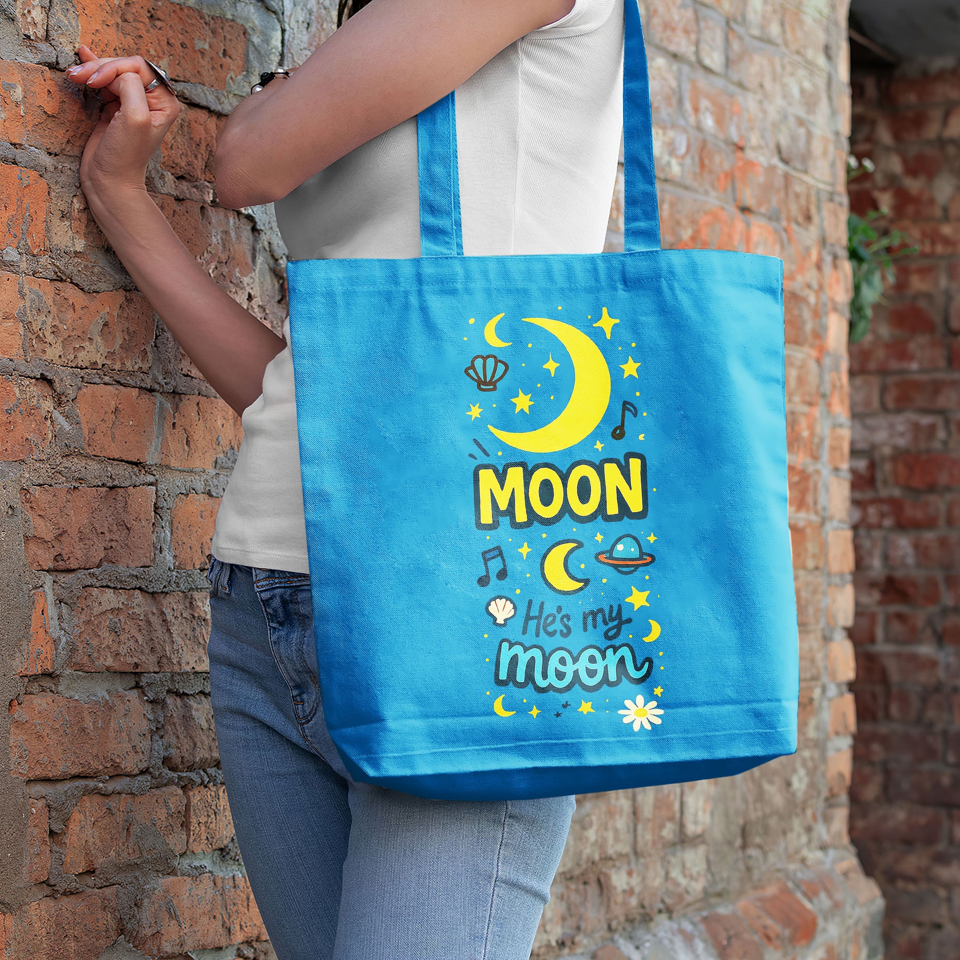 Blue tote bag with yellow moon, stars, and "He's my moon" text on a sandy beach with a straw hat and towel in the background.