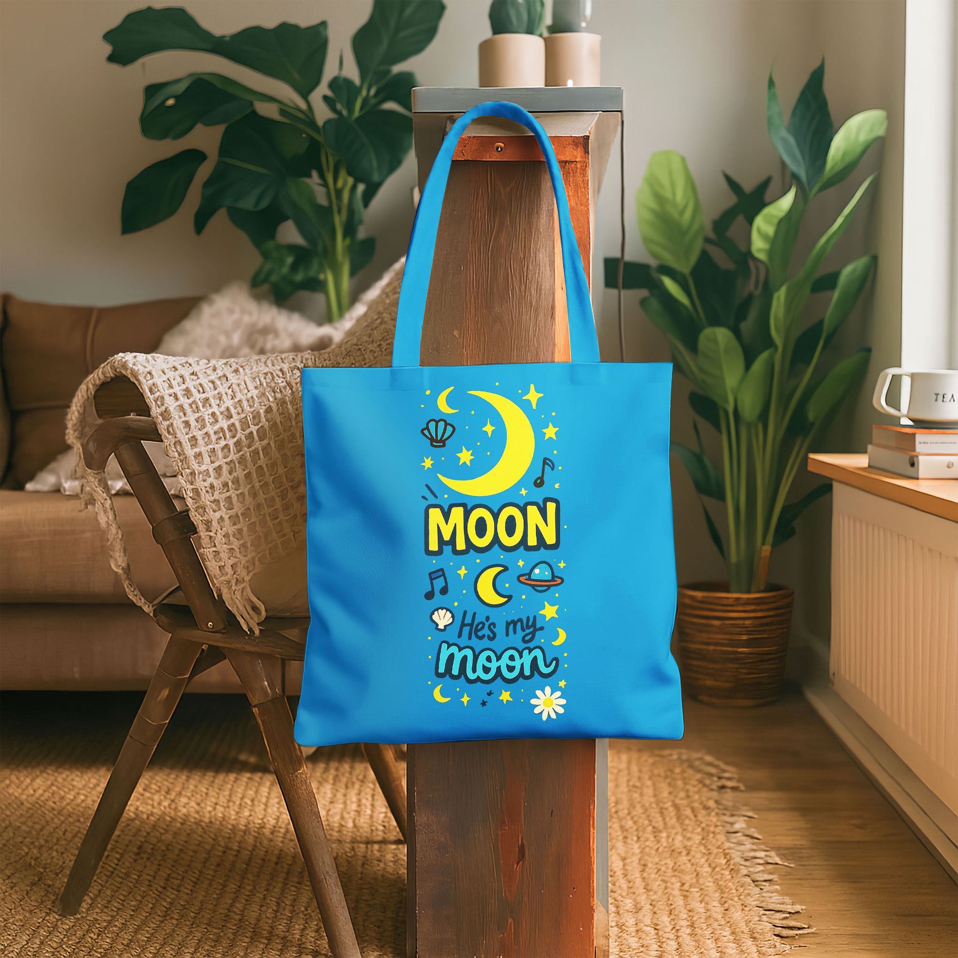 Blue tote bag with yellow moon, stars, and "He's my moon" text on a sandy beach with a straw hat and towel in the background.