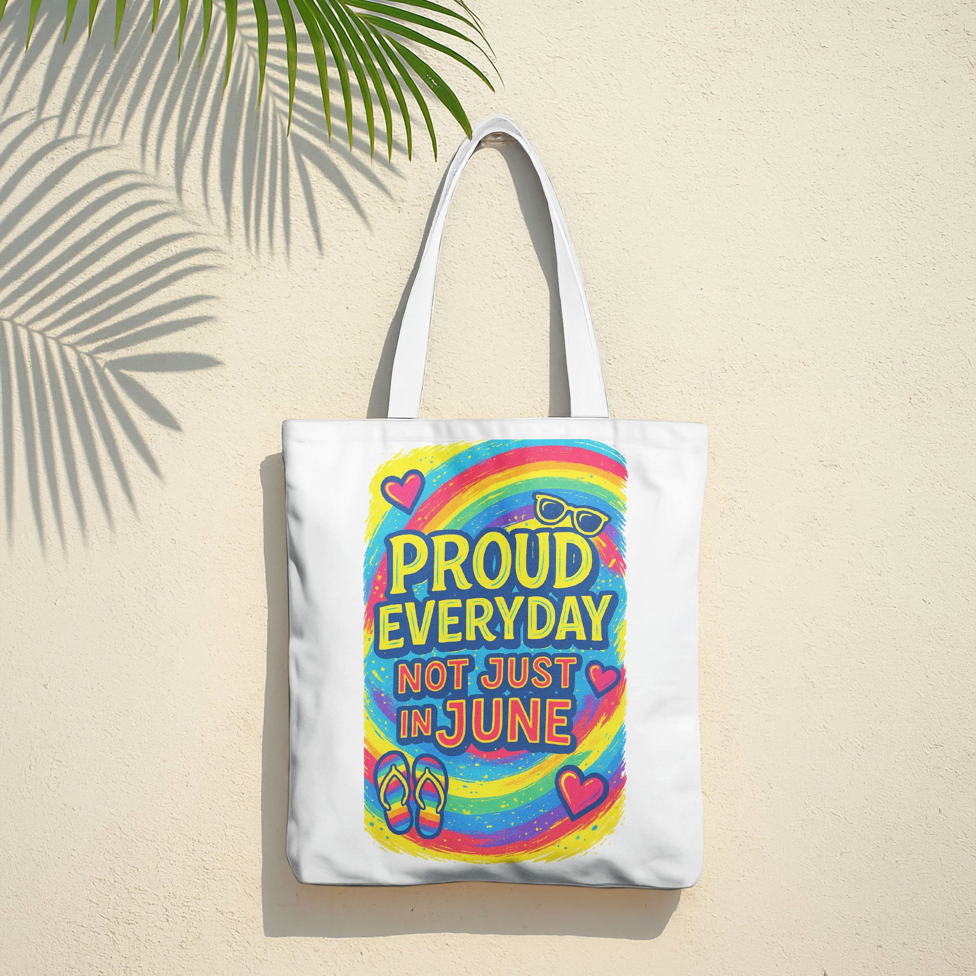 White tote bag with "PROUD EVERYDAY NOT JUST IN JUNE" text, a colorful rainbow, sunglasses, hearts, and flip-flops design, on a sandy beach with the ocean in the background.