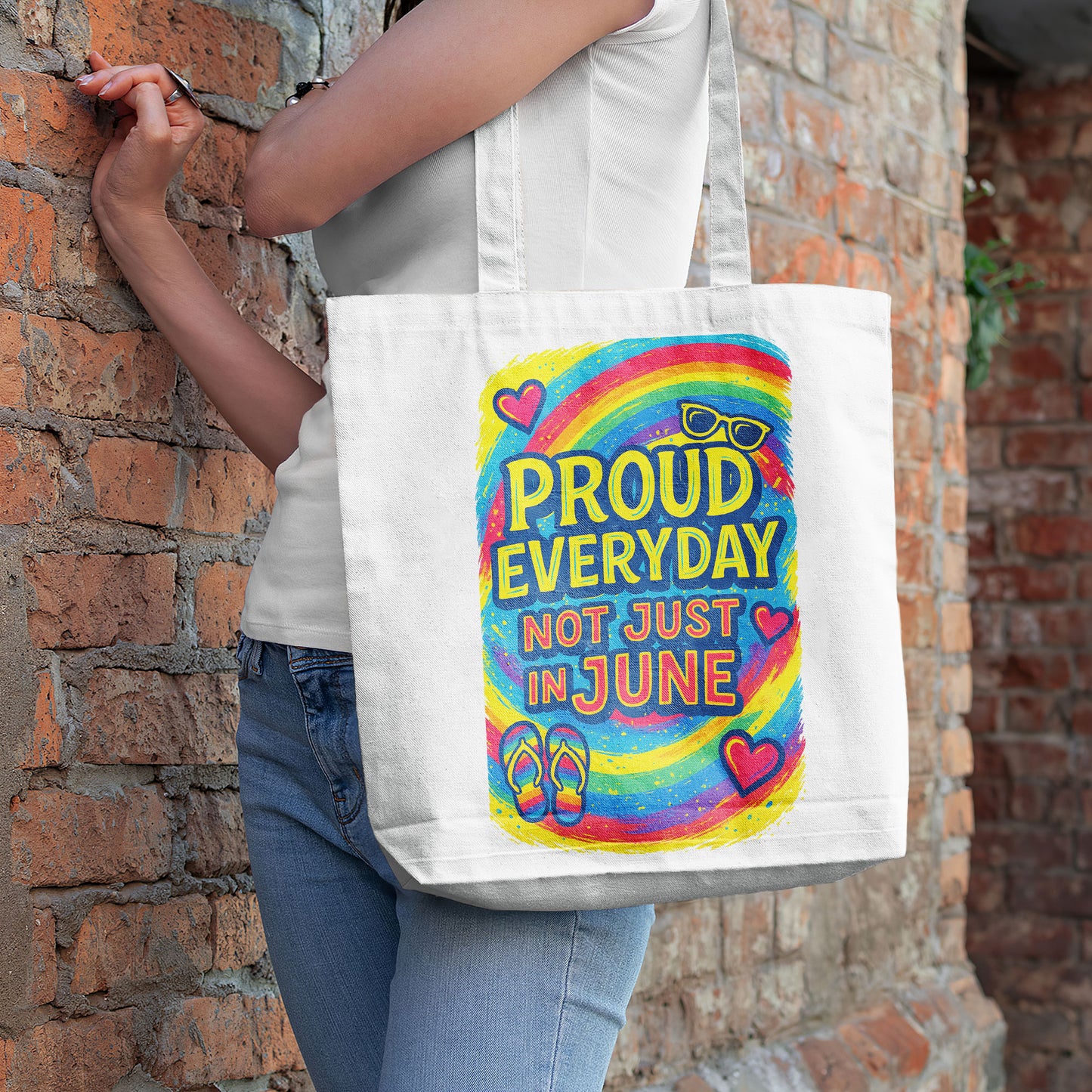 White tote bag with "PROUD EVERYDAY NOT JUST IN JUNE" text, a colorful rainbow, sunglasses, hearts, and flip-flops design, on a sandy beach with the ocean in the background.