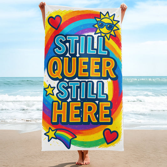 A person holding up a vibrant white beach towel with a swirling rainbow background. The towel features large, bold text "STILL QUEER STILL HERE", along with colorful icons like a sun with sunglasses, hearts, and a rainbow shooting star. The scene is a sunny beach.