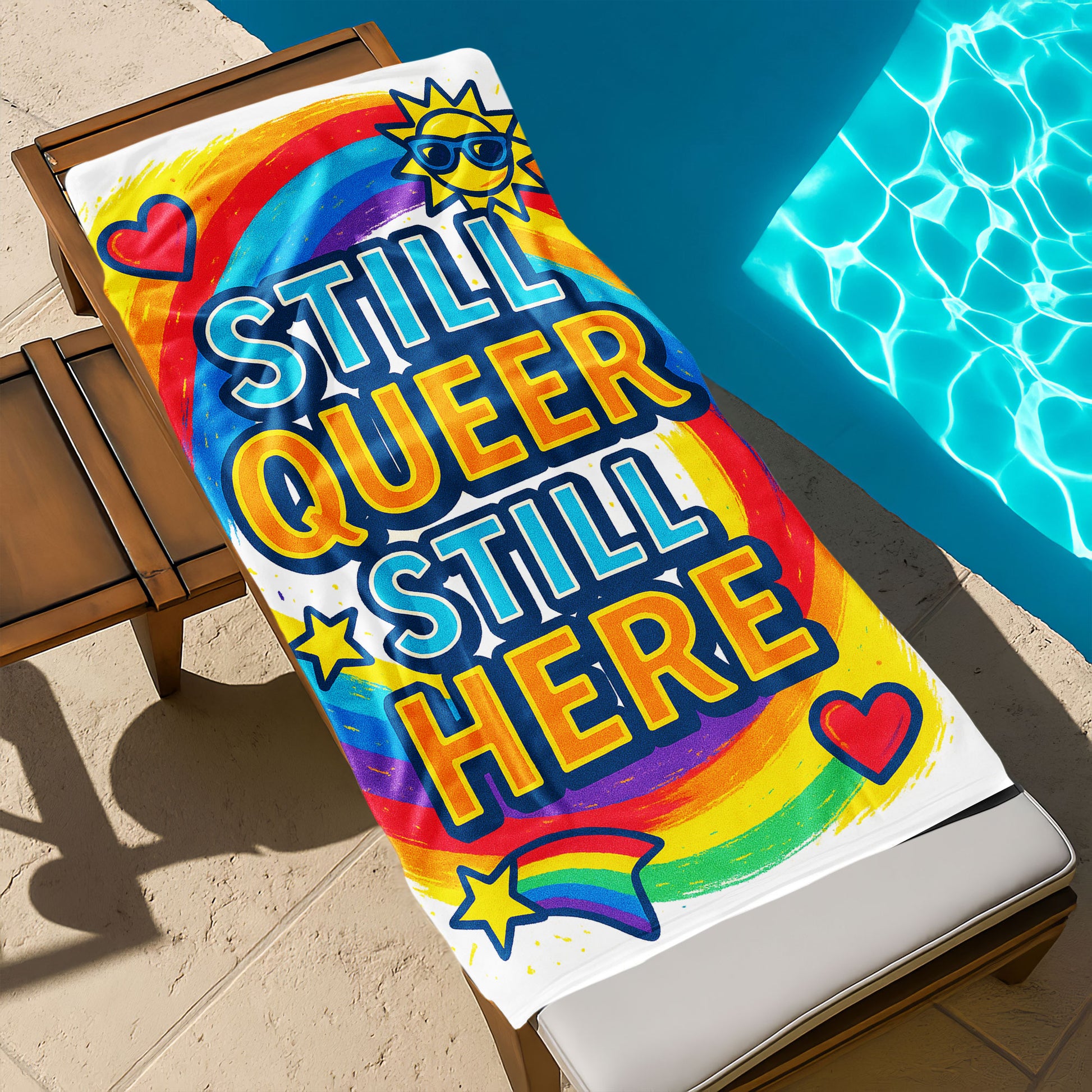 A person holding up a vibrant white beach towel with a swirling rainbow background. The towel features large, bold text "STILL QUEER STILL HERE", along with colorful icons like a sun with sunglasses, hearts, and a rainbow shooting star. The scene is a sunny beach.