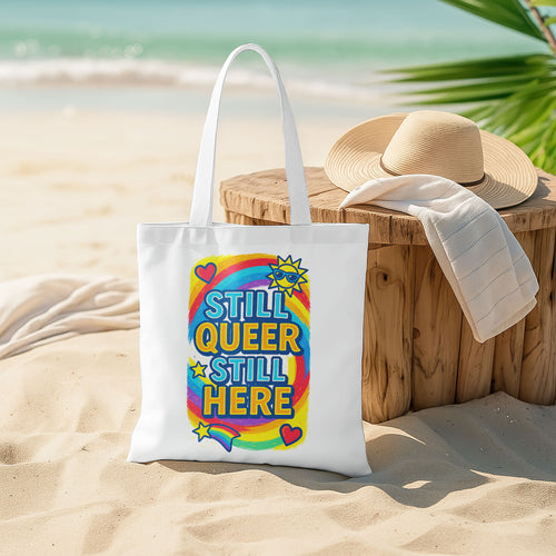 White tote bag with "STILL QUEER STILL HERE" text in colorful letters, a rainbow, stars, hearts, and a sun with sunglasses, on a sandy beach with the ocean in the background.