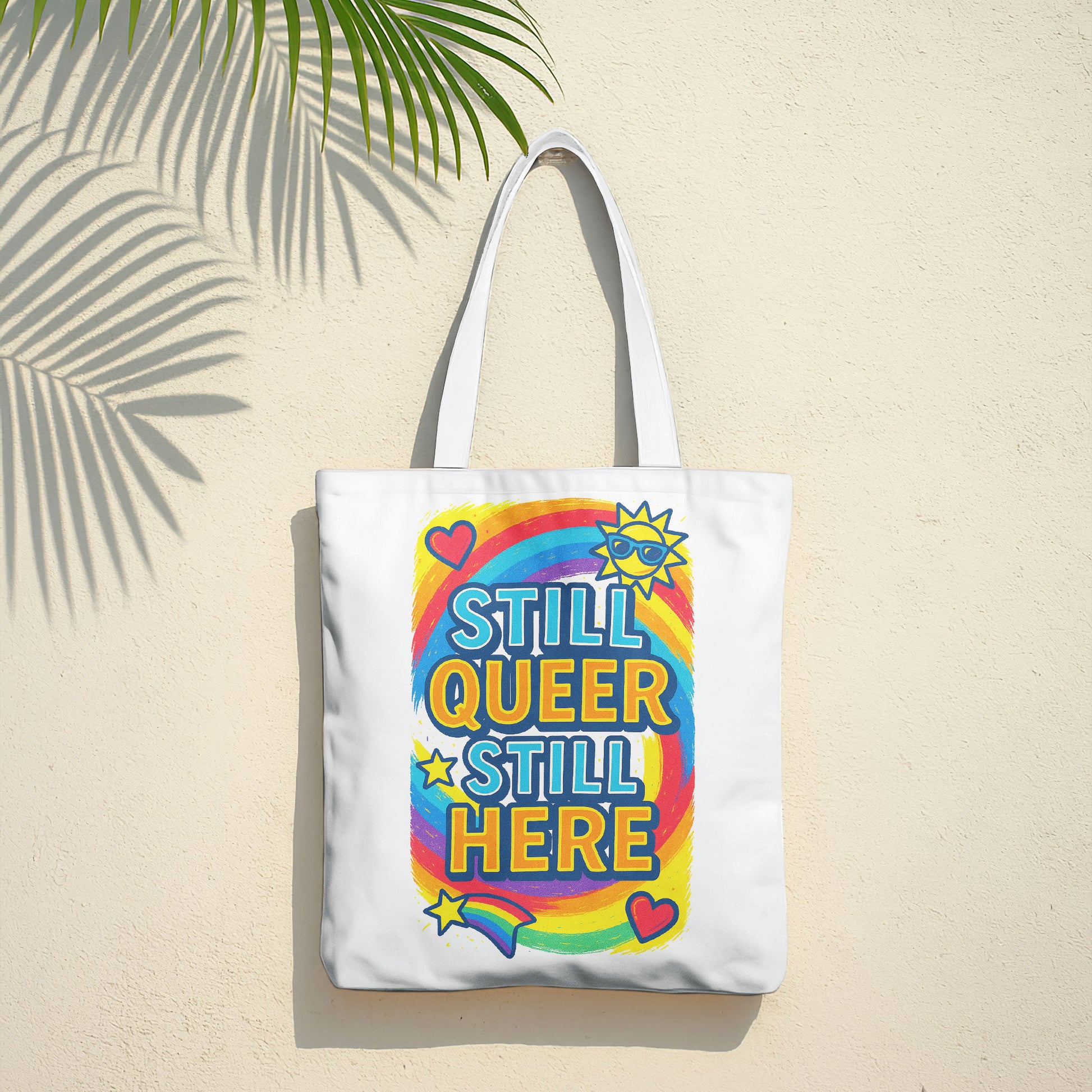 White tote bag with "STILL QUEER STILL HERE" text in colorful letters, a rainbow, stars, hearts, and a sun with sunglasses, on a sandy beach with the ocean in the background.