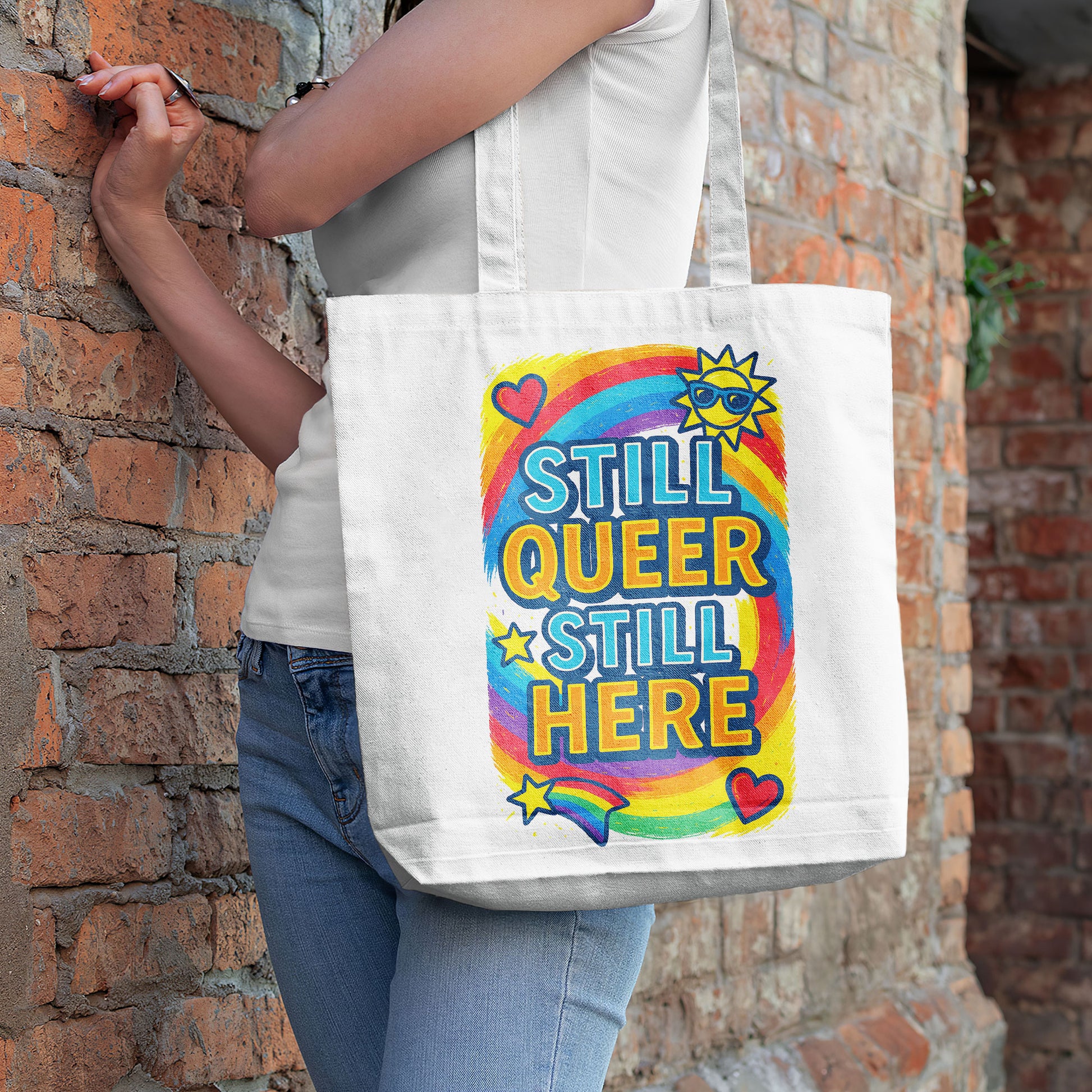 White tote bag with "STILL QUEER STILL HERE" text in colorful letters, a rainbow, stars, hearts, and a sun with sunglasses, on a sandy beach with the ocean in the background.