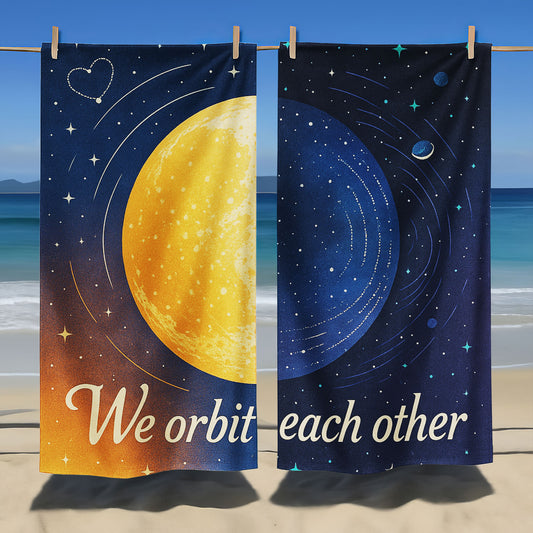 A man and a woman holding up custom beach towels on a beach. One towel shows a large golden planet with stars and "We orbit each other". The other shows a blue planet with orbits and stars, also part of "We orbit each other"