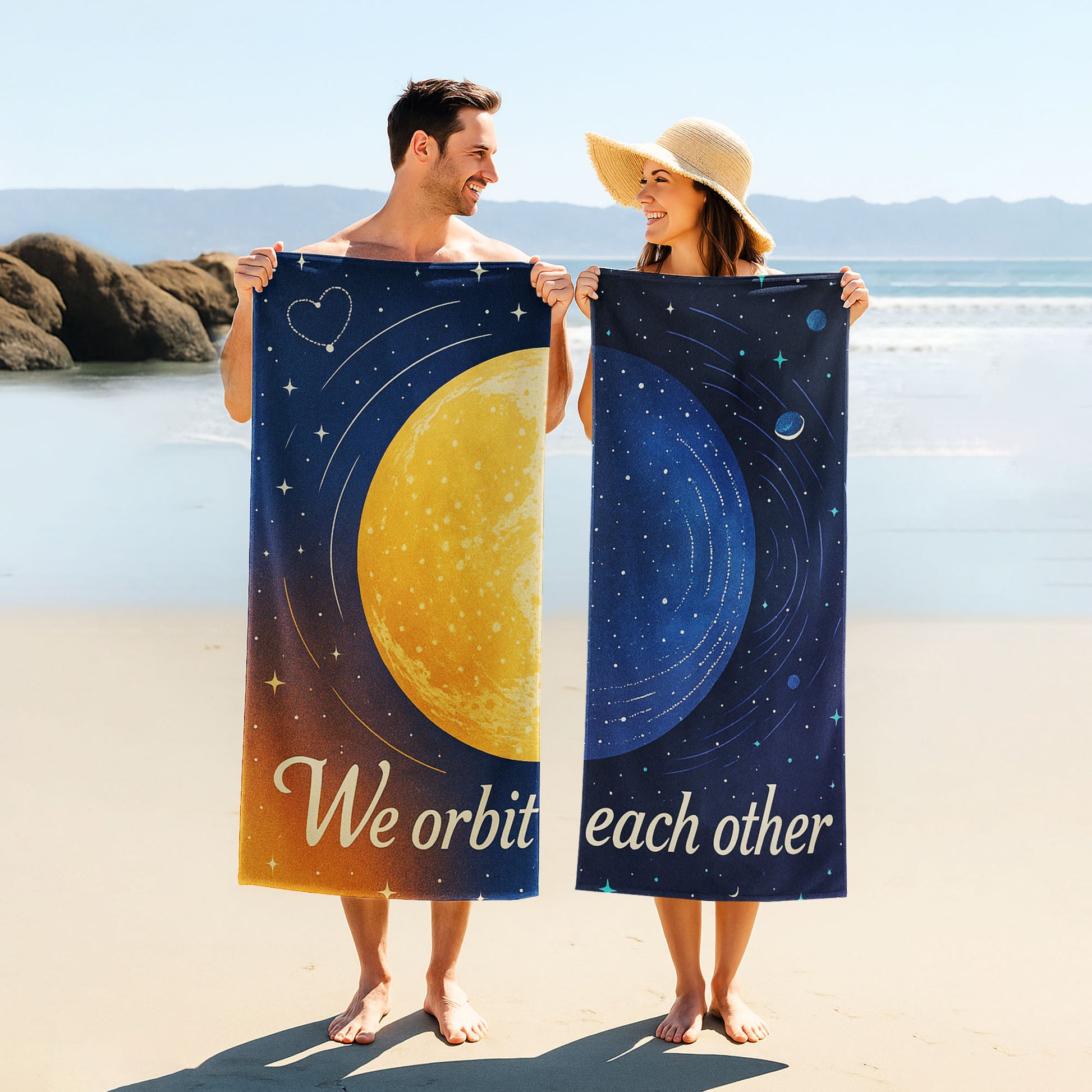  A man and a woman holding up custom beach towels on a beach. One towel shows a large golden planet with stars and "We orbit each other". The other shows a blue planet with orbits and stars, also part of "We orbit each other".