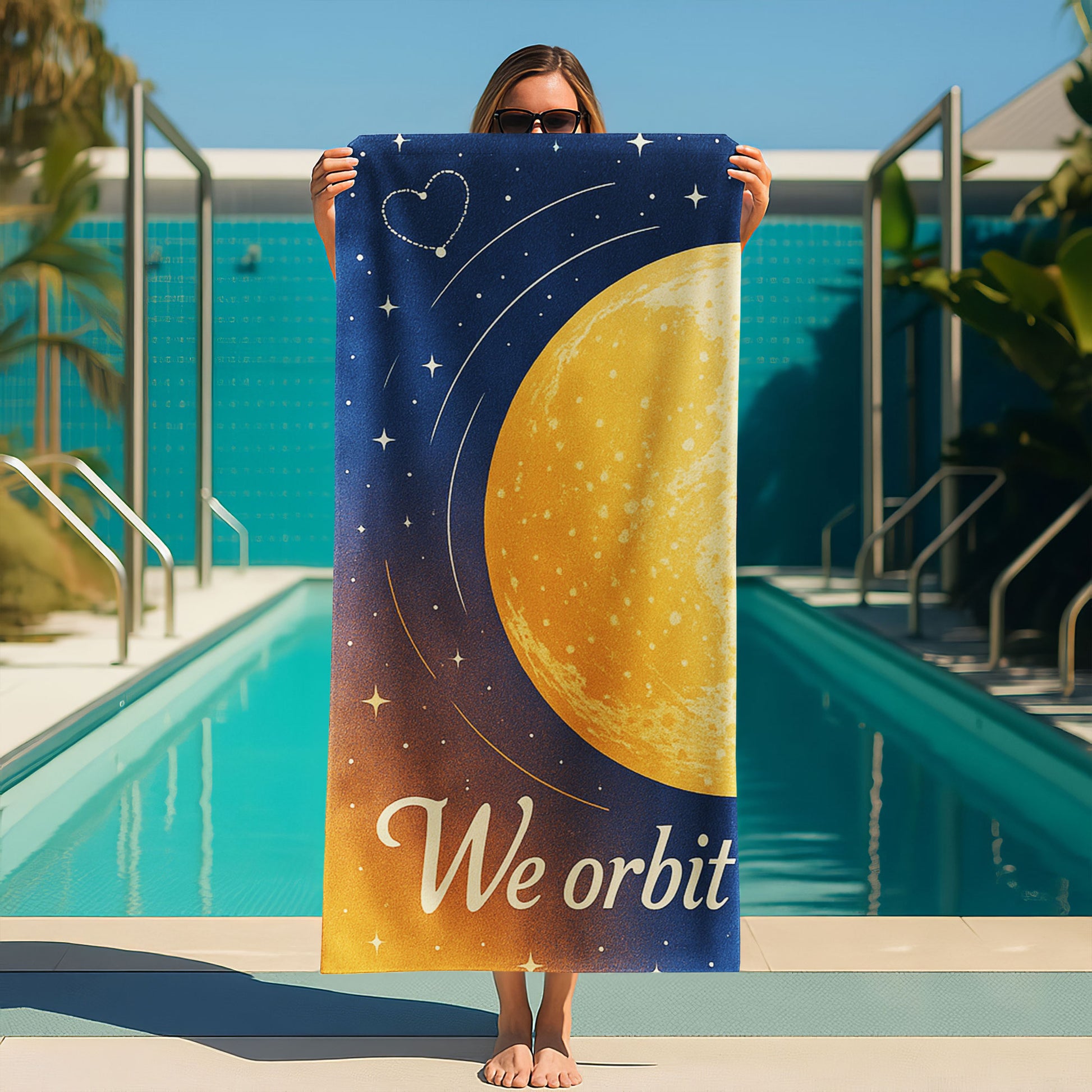  A man and a woman holding up custom beach towels on a beach. One towel shows a large golden planet with stars and "We orbit each other". The other shows a blue planet with orbits and stars, also part of "We orbit each other".